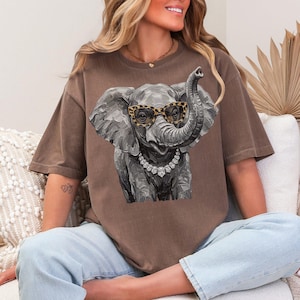 May include: A brown short-sleeve t-shirt featuring a graphic illustration of a gray elephant wearing leopard print glasses and a silver-toned necklace. The elephant is depicted with visible brushstroke textures, and the shirt has a crew neckline.