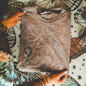 Puede incluir: Una camiseta marr&oacute;n con un dise&ntilde;o gr&aacute;fico blanco que presenta mariposas, setas, flores y corazones. El texto "Just Breathe" est&aacute; escrito en cursiva. La camiseta est&aacute; doblada y sostenida por manos.