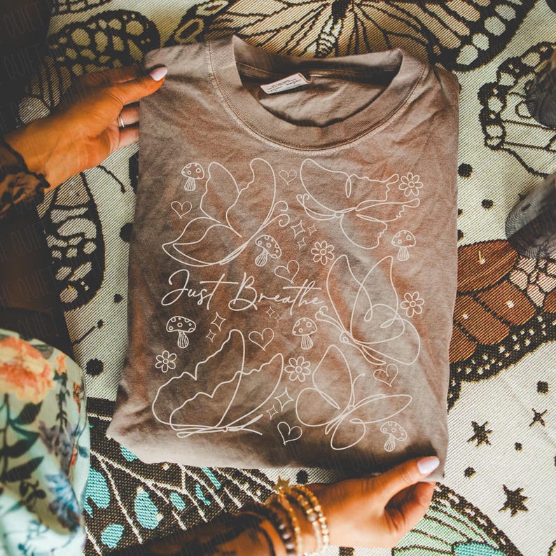 Puede incluir: Una camiseta marr&oacute;n con un dise&ntilde;o gr&aacute;fico blanco que presenta mariposas, setas, flores y corazones. El texto "Just Breathe" est&aacute; escrito en cursiva. La camiseta est&aacute; doblada y sostenida por manos.