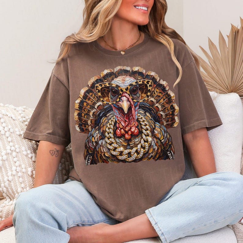 May include: A brown short-sleeve t-shirt featuring a graphic print of a turkey wearing leopard-print glasses and a thick gold chain. The turkey has detailed, colorful feathers in shades of brown, orange, and gold, with a prominent red wattle.