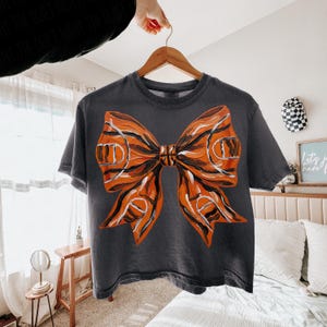 May include: Dark gray t-shirt featuring a large orange and black bow design. The bow has a basketball court pattern and a basketball in the center. The shirt is hanging on a wooden hanger.