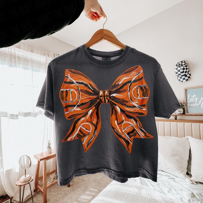 May include: Dark gray t-shirt featuring a large orange and black bow design. The bow has a basketball court pattern and a basketball in the center. The shirt is hanging on a wooden hanger.