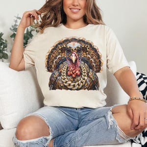 May include: A cream-colored short-sleeve t-shirt featuring a graphic print of a turkey wearing leopard-print sunglasses and a thick gold chain with embedded crystals. The turkey has detailed brown, orange, and black feathers and a red wattle.