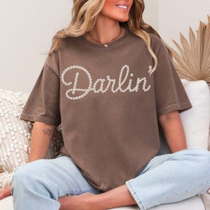 Pu&ograve; includere: T-shirt marrone con la scritta "Darlin'" in perle bianche. La maglietta ha maniche corte e una vestibilit&agrave; ampia. La modella indossa jeans azzurri.