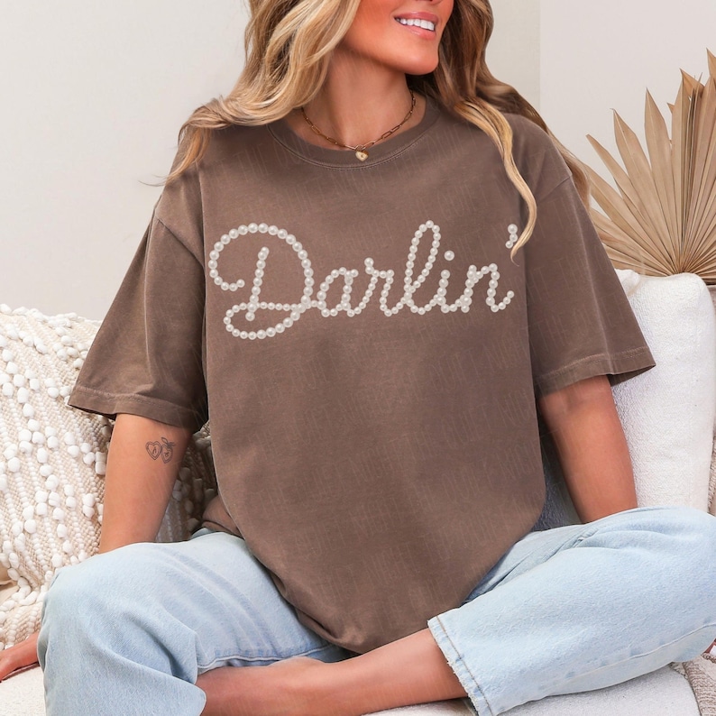 Pu&ograve; includere: T-shirt marrone con la scritta "Darlin'" in perle bianche. La maglietta ha maniche corte e una vestibilit&agrave; ampia. La modella indossa jeans azzurri.