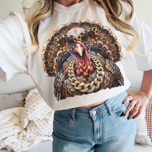 May include: A white t-shirt featuring a graphic print of a turkey wearing leopard print glasses and a thick gold chain with embedded crystals. The turkey has textured, painterly brushstrokes in shades of brown, orange, and red.