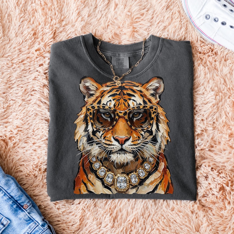 K&ouml;nnte beinhalten: Dunkelgraues T-Shirt mit einer Aquarellillustration eines Tigers, der eine Sonnenbrille mit Leopardenmuster und eine Diamantkette tr&auml;gt. Der Tiger hat orange und schwarze Streifen. Eine goldene Kette ist sichtbar. Das T-Shirt liegt auf einer rosa, flauschigen Oberfl&auml;che.