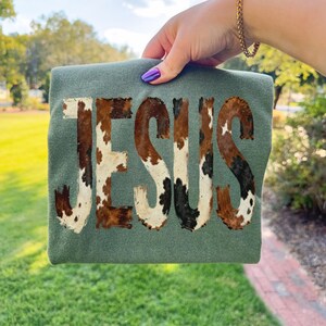 K&ouml;nnte beinhalten: Ein olivgr&uuml;nes T-Shirt mit dem Wort "JESUS" in einem Kuhfellmuster. Die Buchstaben sind braun, wei&szlig; und schwarz. Das Shirt wird von einer Person mit lila Nagellack und einem goldenen Armband gehalten.