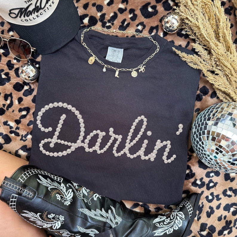 Pu&ograve; includere: T-shirt nero con la scritta "Darlin'" in perle bianche. Sono visibili anche una collana d'oro con ciondoli, un cappello nero, occhiali da sole e stivali da cowboy neri. Lo sfondo ha una stampa leopardata.