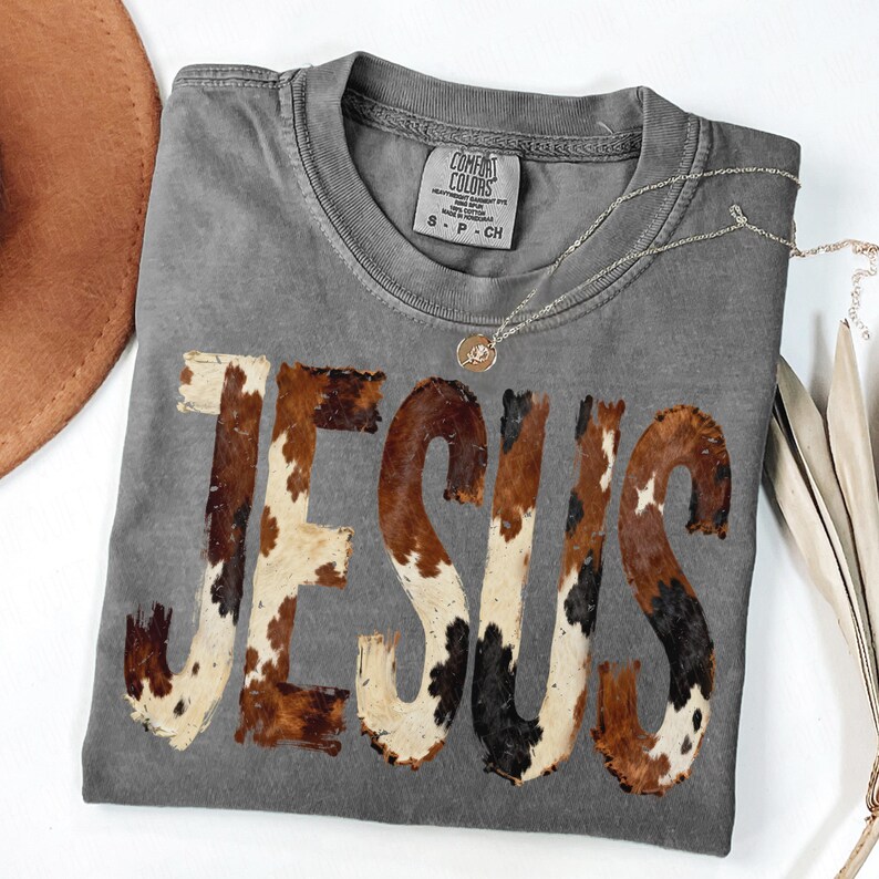 K&ouml;nnte beinhalten: Graues Comfort Colors T-Shirt mit dem Wort "JESUS" im Kuhfellmuster. Eine goldene Halskette mit Anh&auml;nger liegt auf dem Shirt. Ein brauner Hut und getrocknete Pflanzenstiele sind im Hintergrund.