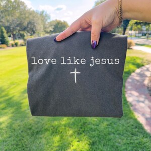 May include: Dark gray t-shirt with the text "love like jesus" and a white cross. The shirt is held up against a green grassy background.