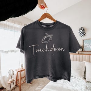 May include: Dark gray t-shirt with the word "Touchdown" in a cursive font, and a football graphic. The shirt is hanging on a wooden hanger.