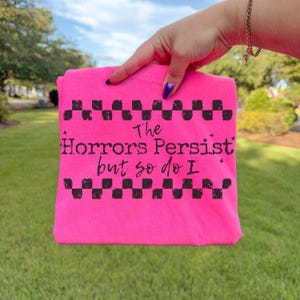 May include: A bright pink t-shirt with the text "The Horrors Persist but so do I" in black. The text is framed by a black checkered pattern. The shirt is held up against a background of green grass and a blue sky.