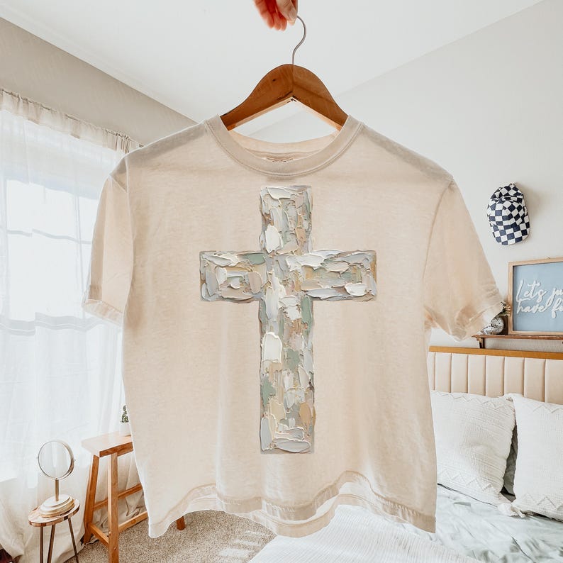 May include: Cream-colored t-shirt featuring a large, textured cross design in shades of white, gray, and beige. The cross appears to be painted with visible brushstrokes. The shirt is hanging on a wooden hanger.