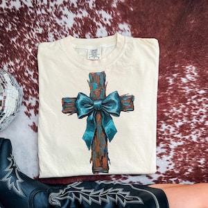 Peut inclure: T-shirt cr&egrave;me avec un motif de croix peinte et un n&oelig;ud bleu sarcelle. La croix est marron et bleu sarcelle, avec un aspect textur&eacute; et rustique. Le t-shirt est de la marque Comfort Colors, taille L-G.