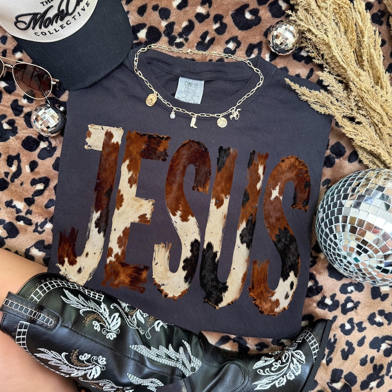 K&ouml;nnte beinhalten: Ein dunkelgraues T-Shirt mit dem Wort "JESUS" im Kuhfellmuster. Das Bild zeigt auch eine schwarze Baseballkappe, Sonnenbrille, eine goldene Halskette, schwarze Cowboy-Stiefel und Discokugeln.