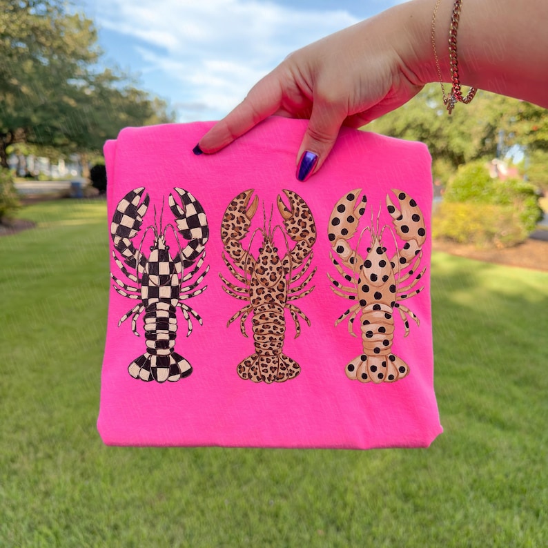 May include: A bright pink t-shirt featuring three lobster designs. The lobsters have unique patterns: a black and white checkerboard, a leopard print, and a tan lobster with black polka dots. The shirt is held up by a hand.