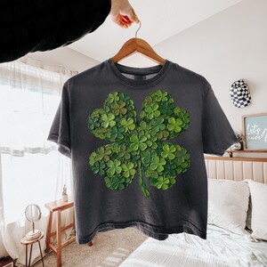 May include: Dark gray t-shirt featuring a large, detailed four-leaf clover design. The clover is composed of many smaller green clovers, creating a textured, layered effect. The shirt is hanging on a wooden hanger.