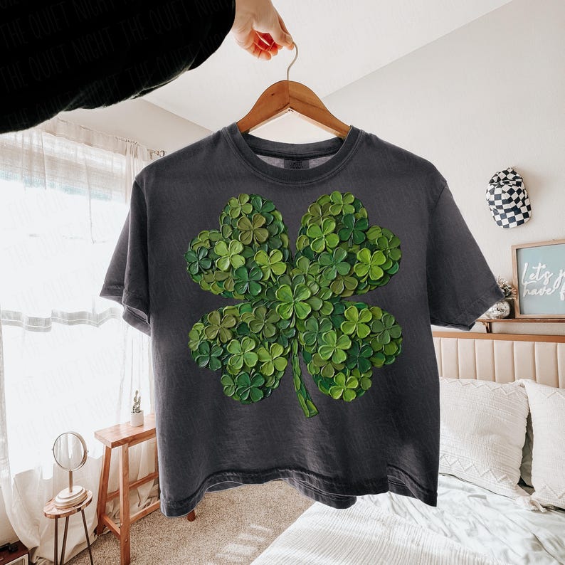 May include: Dark gray t-shirt featuring a large, detailed four-leaf clover design. The clover is composed of many smaller green clovers, creating a textured, layered effect. The shirt is hanging on a wooden hanger.