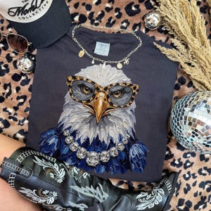 May include: A dark gray t-shirt featuring a graphic of a bald eagle wearing leopard print glasses and a large crystal necklace. The eagle has textured, brushstroke-style feathers in shades of white, gray, and deep blue, set against a dark background.