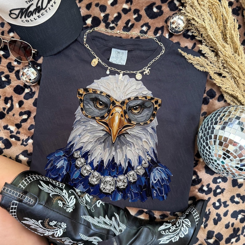 May include: A dark gray t-shirt featuring a graphic of a bald eagle wearing leopard print glasses and a large crystal necklace. The eagle has textured, brushstroke-style feathers in shades of white, gray, and deep blue, set against a dark background.