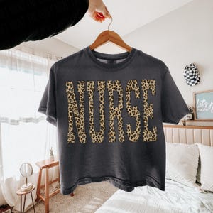 May include: A dark gray t-shirt with the word "NURSE" in large leopard print letters. The shirt is hanging on a wooden hanger. The image is well-lit, showcasing the shirt's design and color.