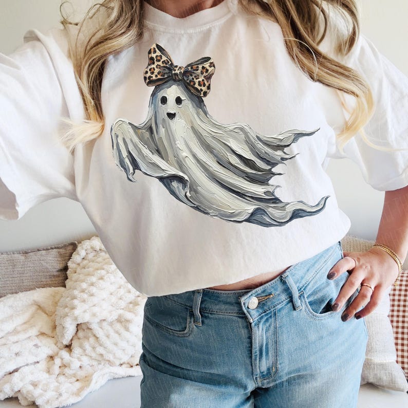 May include: White t-shirt with a ghost design. The ghost is painted in grey tones and wears a leopard print bow. The artwork has a painterly effect, suitable for Halloween or casual wear.