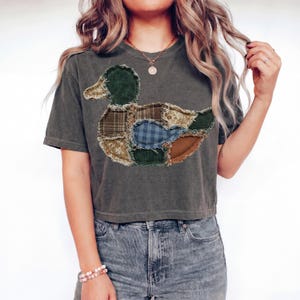 May include: A charcoal gray short-sleeve t-shirt featuring a large central graphic of a duck composed of various fabric patches. The duck design includes textures like corduroy, plaid, and floral patterns in shades of green, blue, and brown.