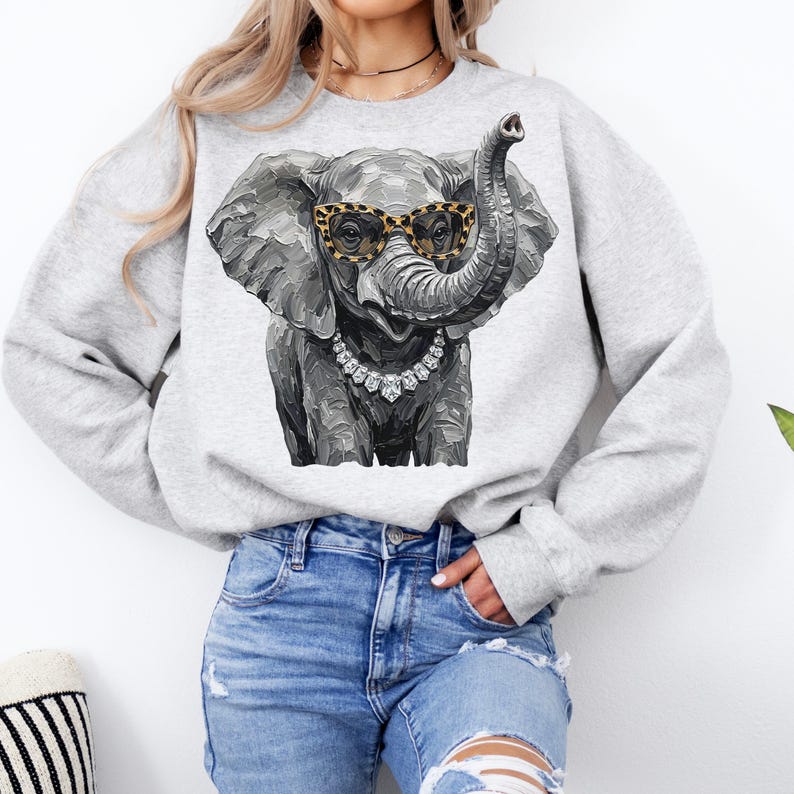 May include: A light gray crewneck sweatshirt featuring a graphic print of an elephant wearing leopard print glasses and a silver-toned crystal necklace. The elephant illustration has a textured, painterly style with shades of gray and black.
