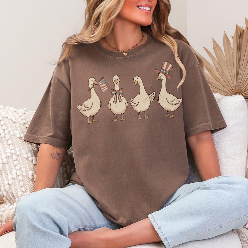 May include: Brown t-shirt featuring a whimsical illustration of four geese. One goose holds an American flag, another wears a bow, and the last wears a patriotic top hat. The geese are in a row, adding a touch of fun to the design.