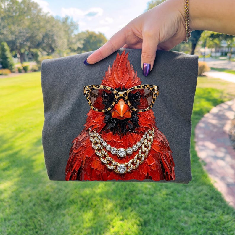 May include: A dark gray t-shirt featuring a vibrant red cardinal wearing leopard print sunglasses and a gold chain necklace with diamond accents. The bird has a detailed, textured appearance.