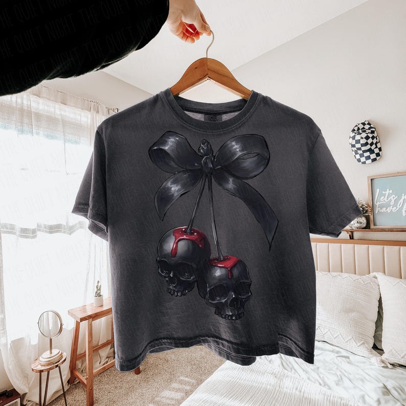 May include: Dark grey t-shirt with a graphic of two black skulls with red accents, styled as cherries, tied with a black bow. The shirt is hanging on a wooden hanger.
