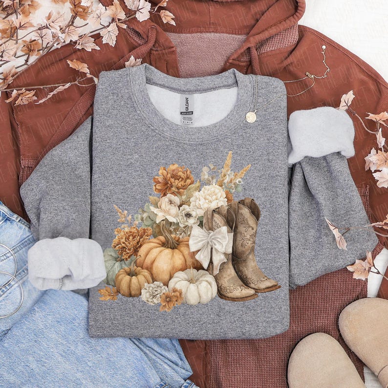 May include: A gray sweatshirt featuring a fall-themed design. The graphic includes pumpkins, flowers, and cowboy boots. The sweatshirt is surrounded by a rust-colored hoodie and denim jeans, creating a cozy, autumnal aesthetic.