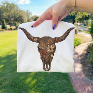 May include: A white t-shirt featuring a detailed painting of a bull skull adorned with floral accents in shades of brown, beige, and cream. The artwork is centered on the shirt, showcasing a rustic, artistic design. The shirt is held up against a green lawn.