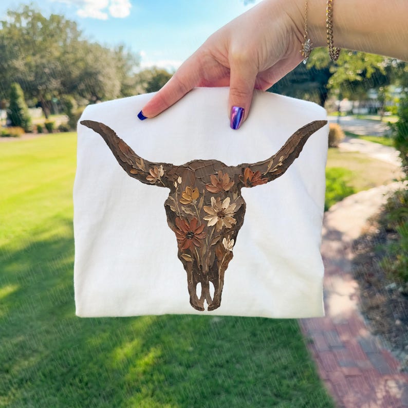 May include: A white t-shirt featuring a detailed painting of a bull skull adorned with floral accents in shades of brown, beige, and cream. The artwork is centered on the shirt, showcasing a rustic, artistic design. The shirt is held up against a green lawn.