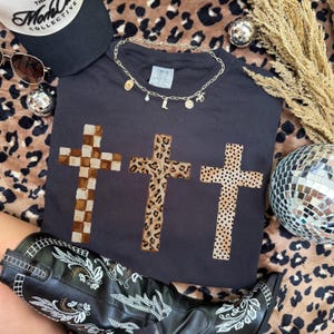 May include: A black t-shirt featuring three crosses with different patterns: checkered, leopard print, and dotted. A gold necklace with charms rests on the shirt. Accessories include a black cap, sunglasses, and a disco ball.