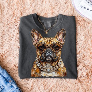 May include: Dark gray t-shirt featuring a detailed illustration of a French Bulldog wearing leopard print glasses and a diamond-studded chain. The dog's fur is a mix of brown and tan. A gold chain necklace is also visible. The shirt is on a pink fluffy surface.