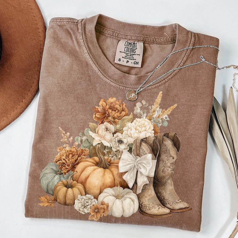 May include: A brown Comfort Colors t-shirt featuring a fall-themed graphic. The design includes pumpkins, flowers, and cowboy boots. A silver necklace is draped across the shirt. The shirt has a crew neck and short sleeves.