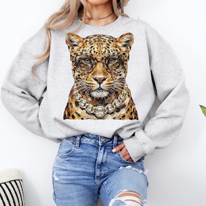 Puede incluir: Una sudadera gris claro con una ilustraci&oacute;n de leopardo que lleva gafas y un collar de diamantes. El leopardo tiene un pelaje manchado en tonos marrones y beige. Las gafas son marrones y el collar tiene grandes gemas cuadradas.