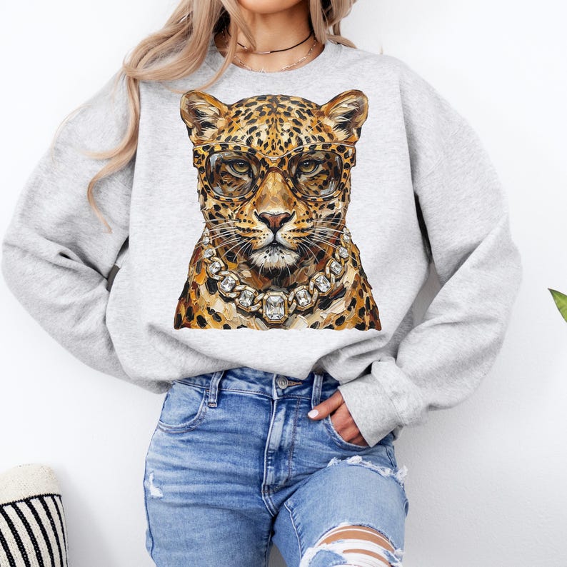 Puede incluir: Una sudadera gris claro con una ilustraci&oacute;n de leopardo que lleva gafas y un collar de diamantes. El leopardo tiene un pelaje manchado en tonos marrones y beige. Las gafas son marrones y el collar tiene grandes gemas cuadradas.
