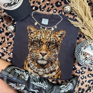 Puede incluir: Una camiseta negra con un leopardo con gafas y un collar de diamantes. La imagen incluye una gorra de b&eacute;isbol negra, gafas de sol, un collar dorado, una bola de discoteca y botas de vaquero negras con bordados blancos, todo sobre un fondo de estampado de leopardo.