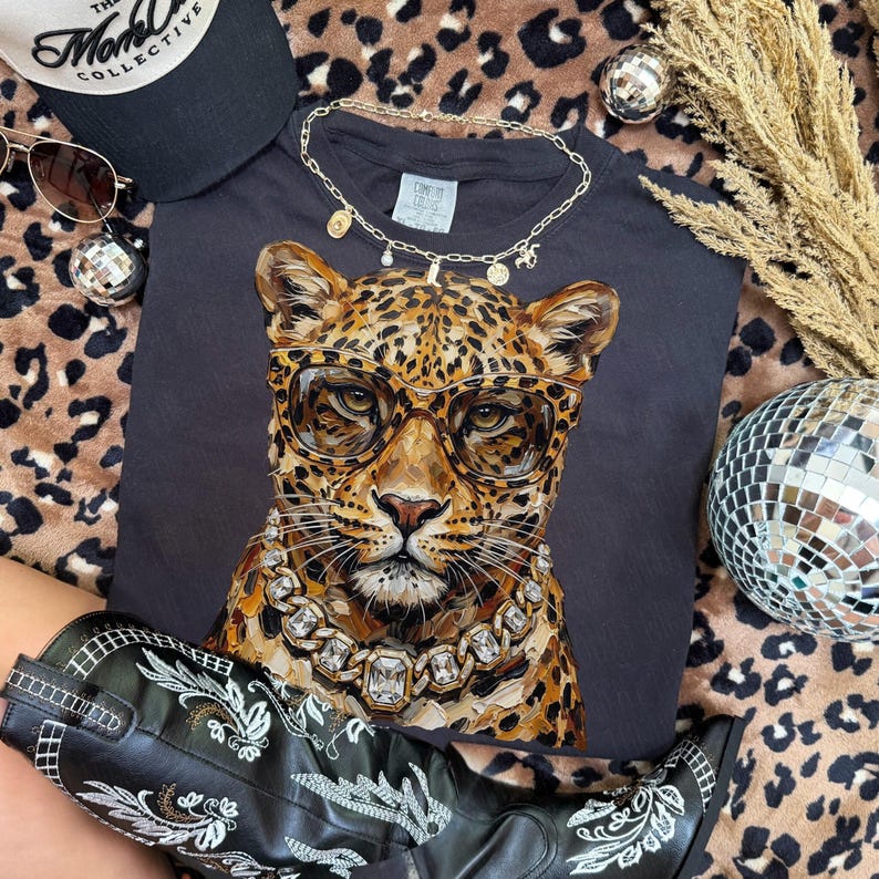 Puede incluir: Una camiseta negra con un leopardo con gafas y un collar de diamantes. La imagen incluye una gorra de b&eacute;isbol negra, gafas de sol, un collar dorado, una bola de discoteca y botas de vaquero negras con bordados blancos, todo sobre un fondo de estampado de leopardo.
