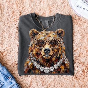 May include: Dark grey t-shirt featuring a detailed illustration of a bear wearing leopard print glasses and a diamond necklace. The bear's fur is rendered in shades of brown and gold. A gold chain is also visible. The shirt is folded on a pink surface.