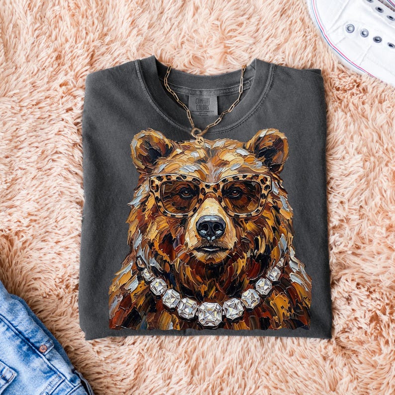 May include: Dark grey t-shirt featuring a detailed illustration of a bear wearing leopard print glasses and a diamond necklace. The bear's fur is rendered in shades of brown and gold. A gold chain is also visible. The shirt is folded on a pink surface.