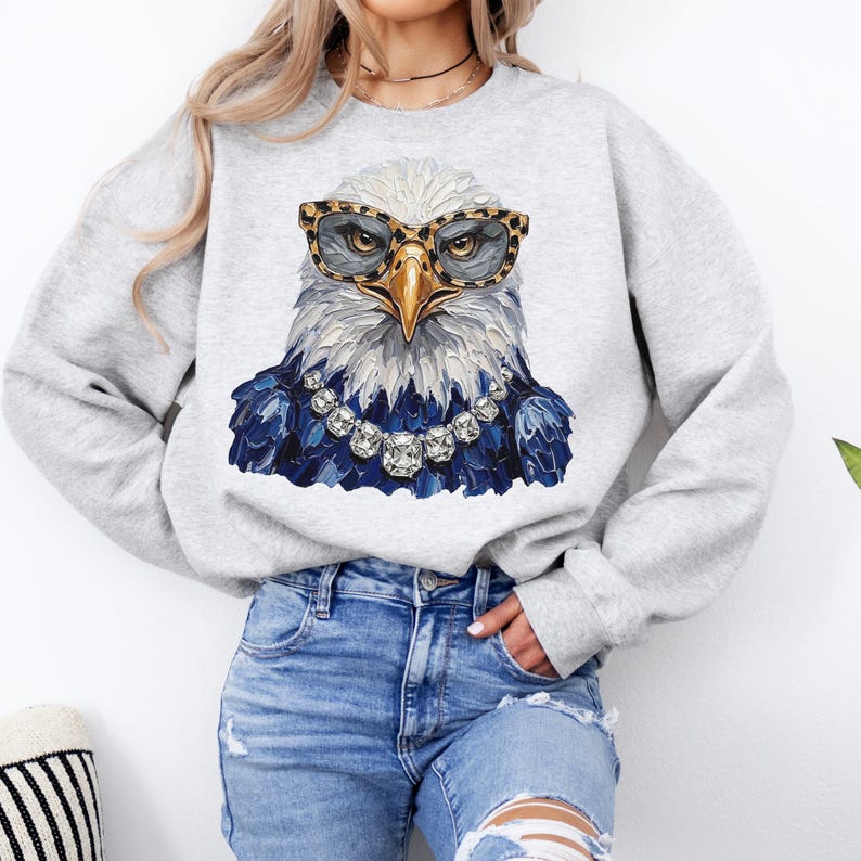 May include: A light gray crewneck sweatshirt featuring a central graphic of a bald eagle wearing leopard print glasses and a large, multi-faceted crystal necklace. The eagle is rendered in a textured, painterly style with blue plumage.