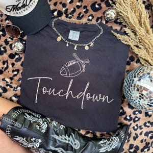 May include: Black t-shirt with a football graphic and the word "Touchdown" in cursive. A gold necklace with charms, a black baseball cap, sunglasses, and black cowboy boots are also visible. The background has a leopard print.