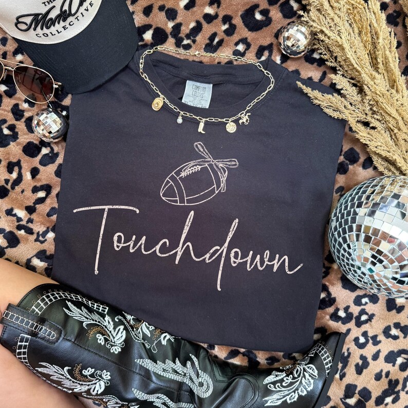 May include: Black t-shirt with a football graphic and the word "Touchdown" in cursive. A gold necklace with charms, a black baseball cap, sunglasses, and black cowboy boots are also visible. The background has a leopard print.