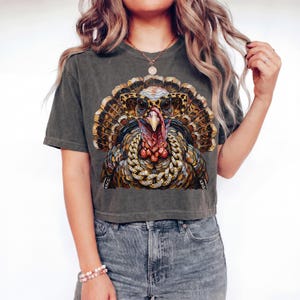May include: A charcoal gray short-sleeve t-shirt featuring a graphic print of a turkey wearing leopard-print glasses and a thick gold chain. The turkey has detailed brown, tan, and orange feathers, a red wattle, and a prominent beak.
