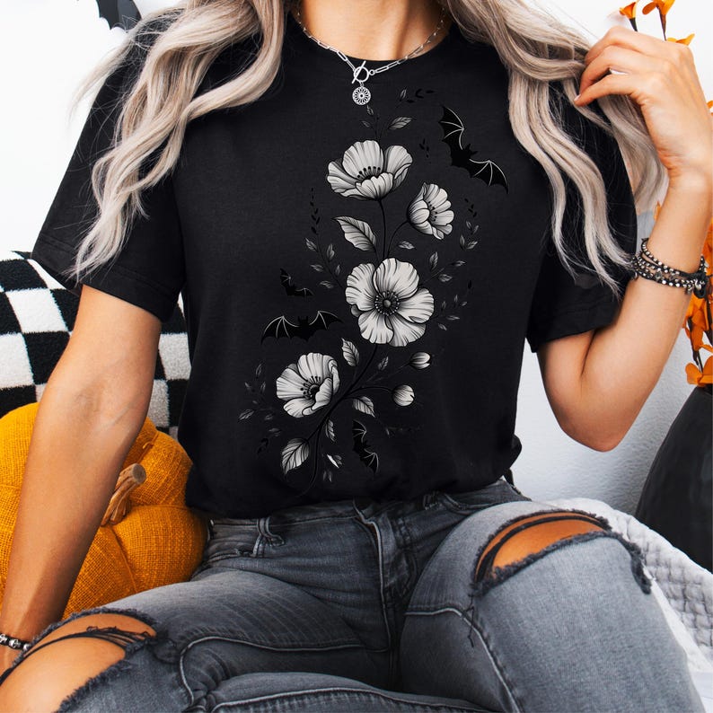 May include: Black t-shirt featuring a floral design with white flowers and leaves, accented by small bat silhouettes. The shirt has a crew neck and short sleeves. The design is centered on the front of the shirt.