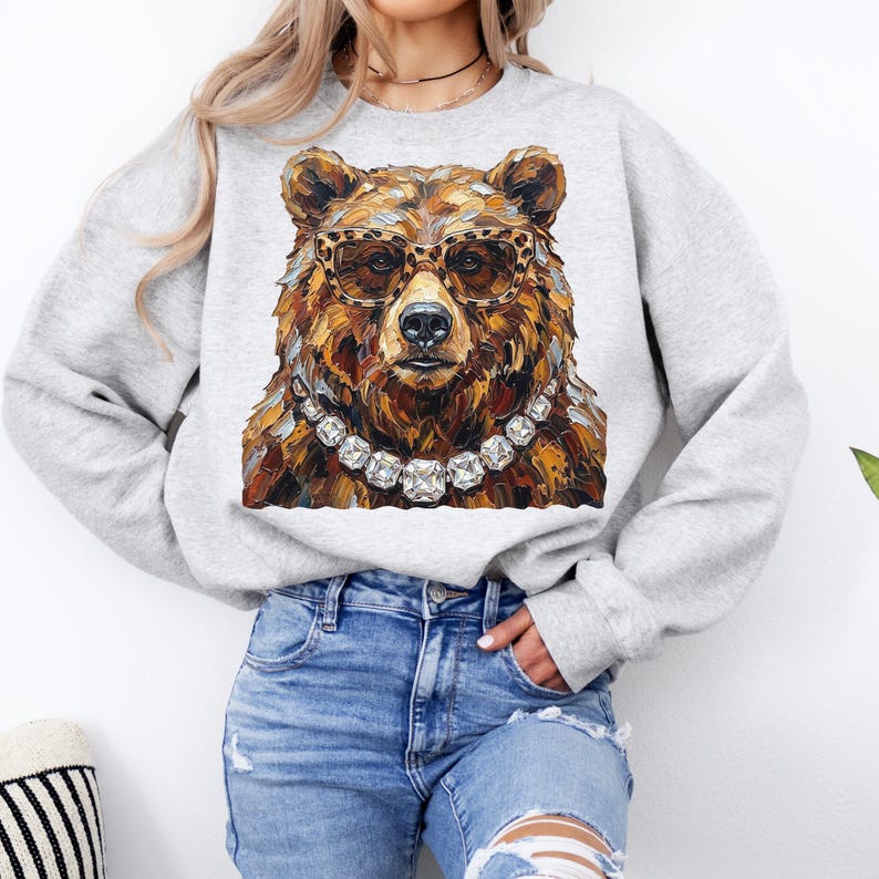 May include: A light gray sweatshirt featuring a detailed illustration of a bear wearing leopard print glasses and a diamond necklace. The bear's fur is rendered in shades of brown and gold. The sweatshirt is a crew neck style.